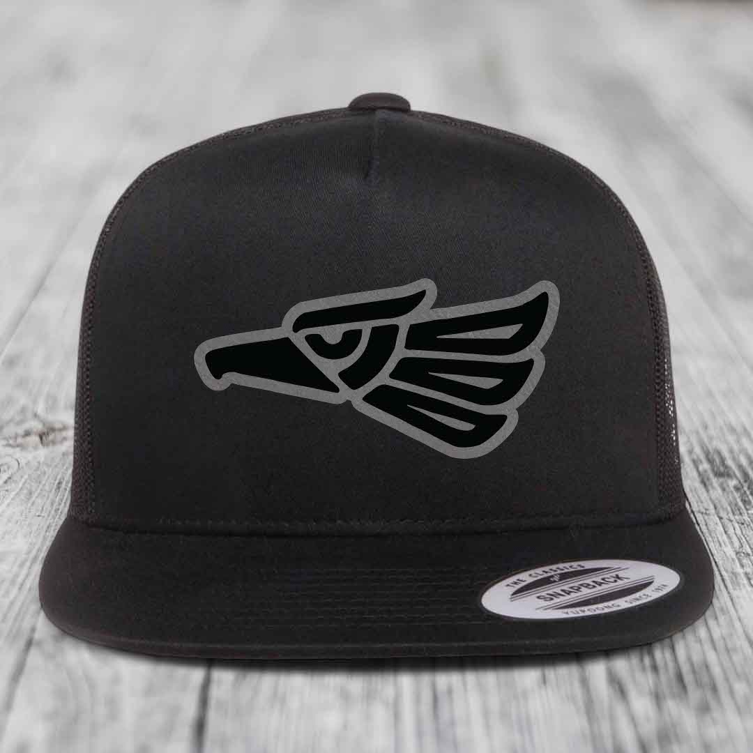 Black snapback trucker hat with Aztec-style eagle patch on front, resting on wooden surface.