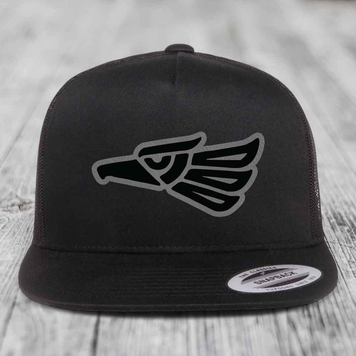 Black snapback trucker hat with Aztec-style eagle patch on front, resting on wooden surface.