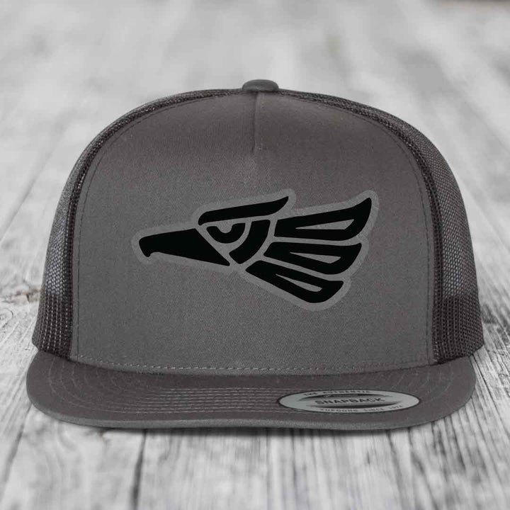 Grey trucker hat with black Aztec-style eagle patch on front, flat brim, mesh back.