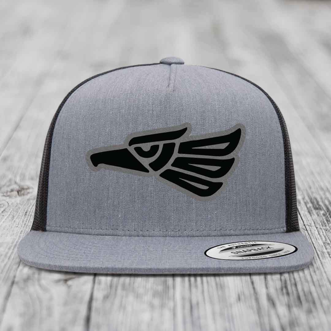 Grey trucker hat with black Aztec-style eagle patch on front, black mesh back, and Yupoong brand label.