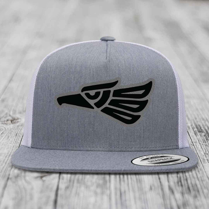 Grey trucker hat with black Aztec eagle patch on front, white mesh back, flat brim.