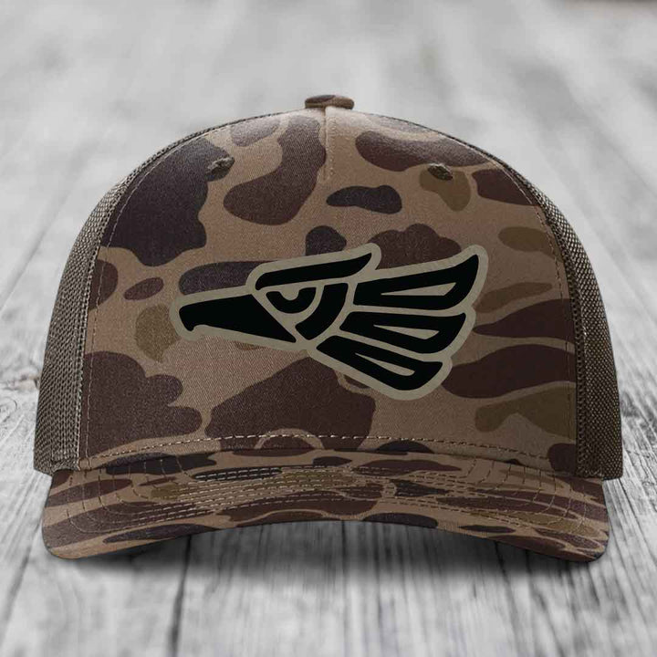 Camouflage trucker hat with black eagle patch on front, sitting on wooden surface.
