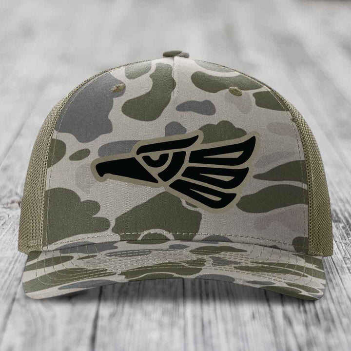 Camouflage trucker hat with black Aztec eagle patch on front.