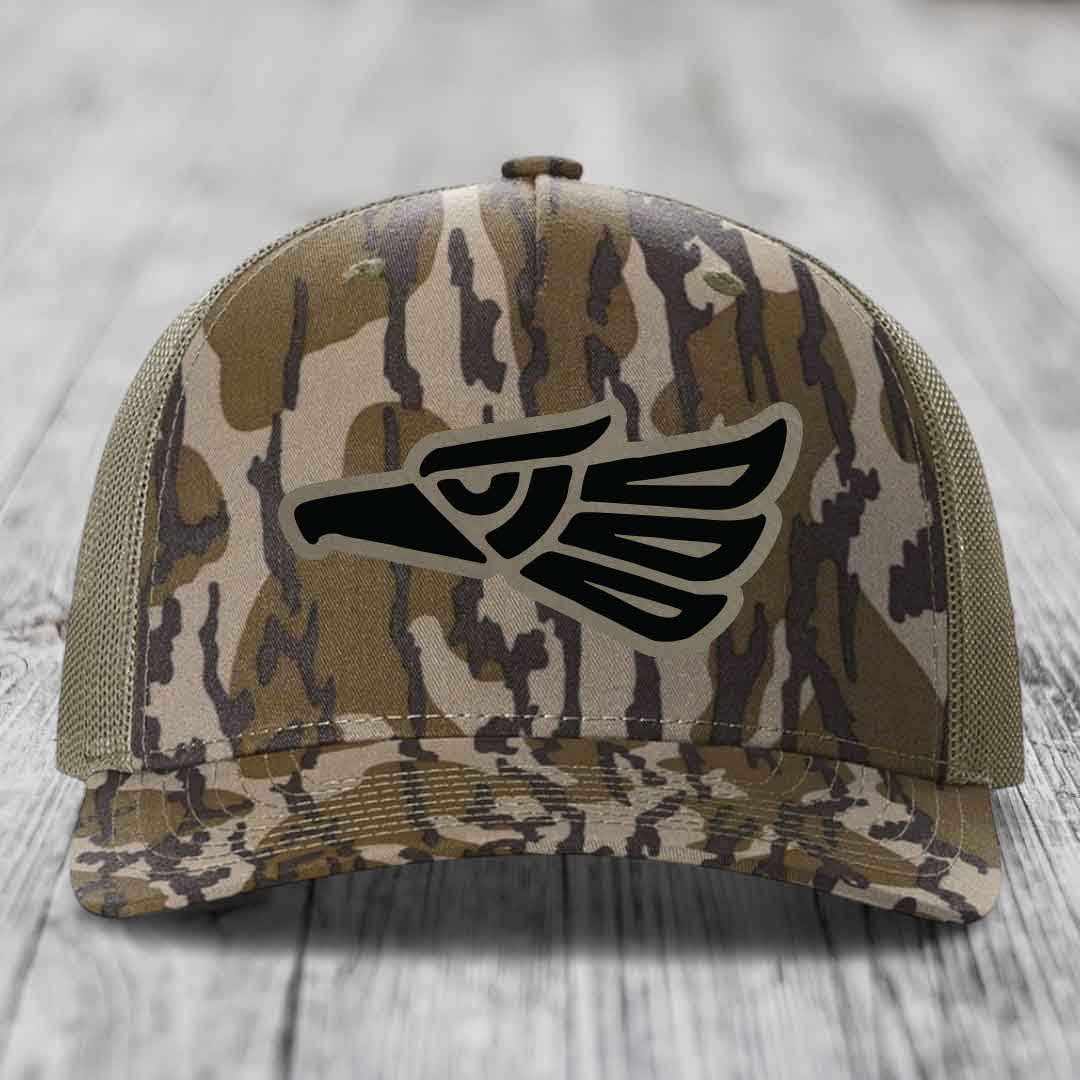 Camouflage trucker hat with black eagle patch on front, mesh back, wooden surface background.