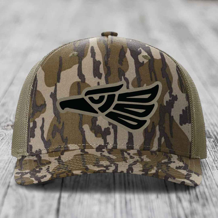 Camouflage trucker hat with black eagle patch on front, mesh back, wooden surface background.