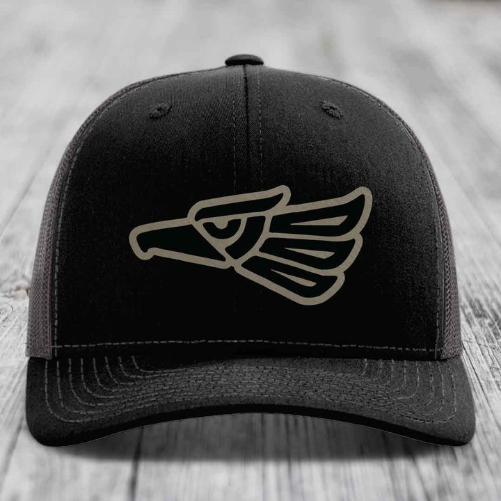 Black trucker hat with embroidered Aztec eagle patch on front, placed on wooden surface.
