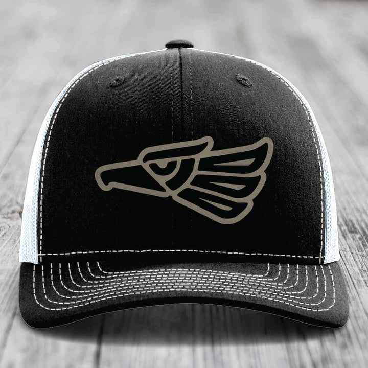 Black trucker hat with white mesh back and stitching, featuring a grey eagle head patch on front.