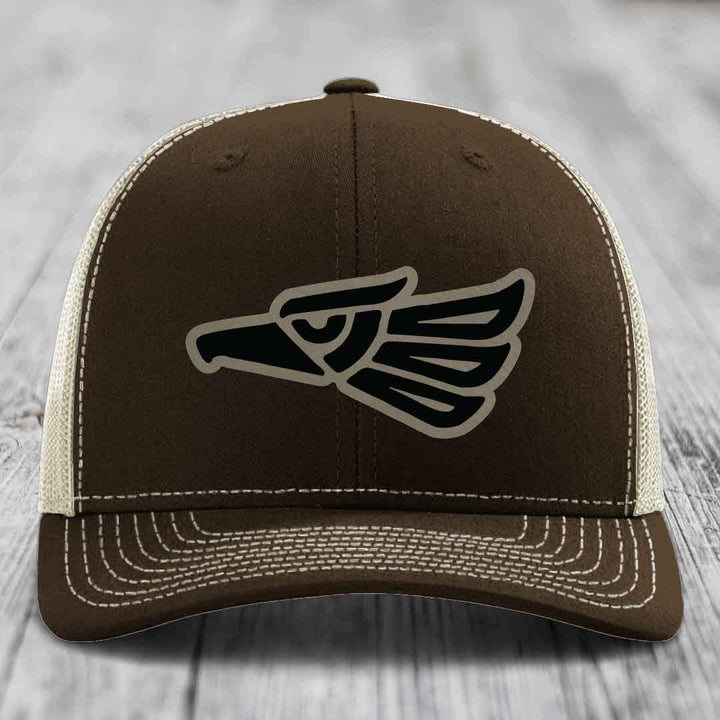 Brown trucker hat with black and tan Aztec eagle patch on front, white stitching details.