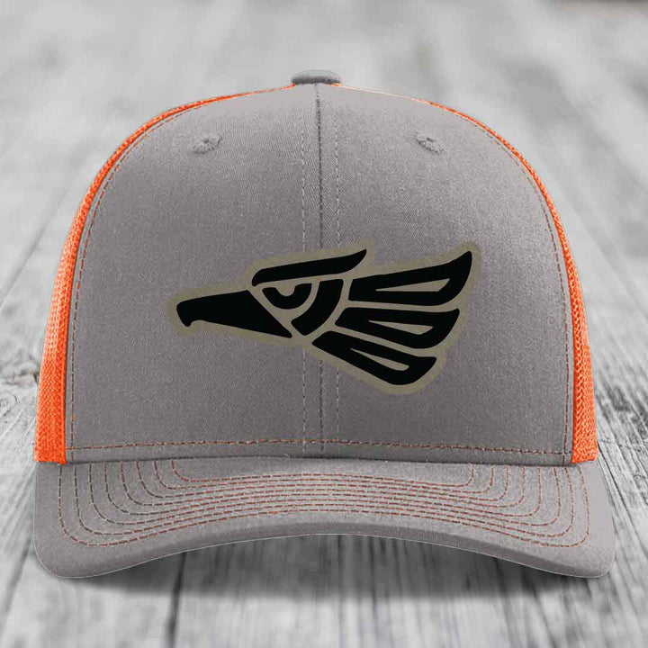 Grey trucker hat with orange mesh back and black Aztec eagle patch on front.