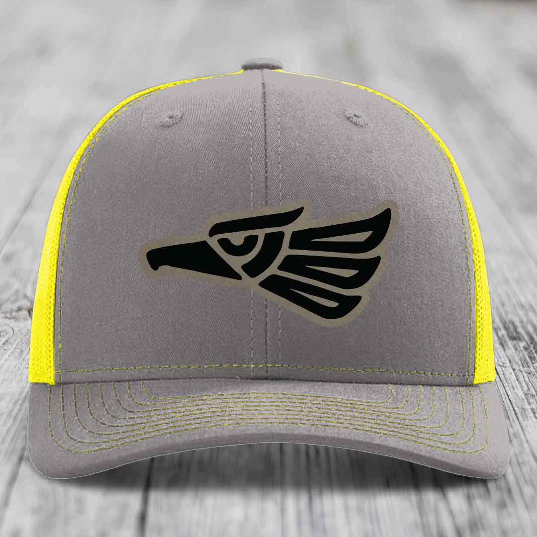 Grey trucker hat with black Aztec eagle patch and yellow mesh sides and stitching.