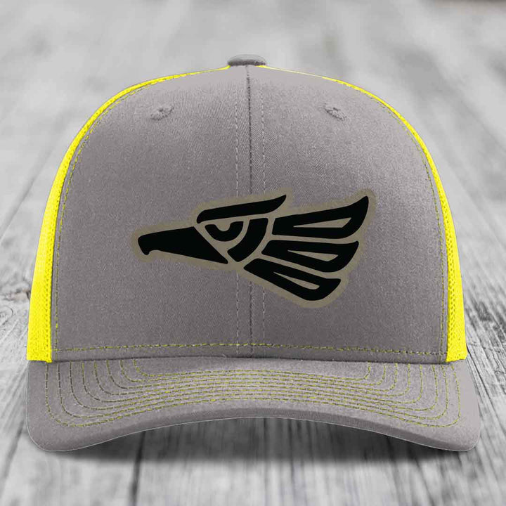 Grey trucker hat with black Aztec eagle patch and yellow mesh sides and stitching.