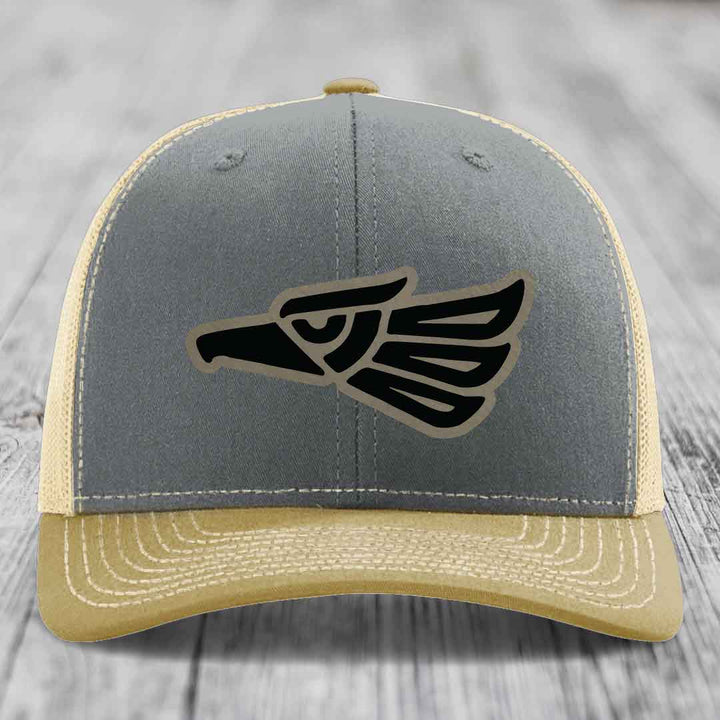 Grey and beige trucker hat with black Aztec eagle leather patch on front.