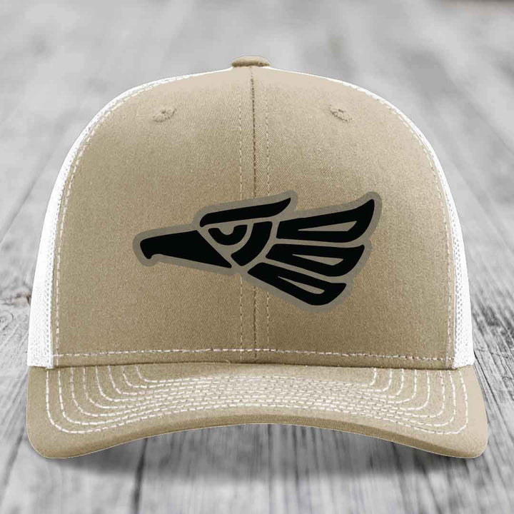 Tan trucker hat with black Aztec eagle patch on front, white mesh back and stitching.