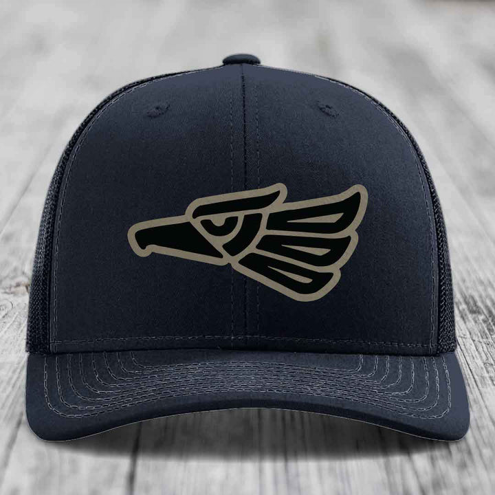 Black trucker hat with Aztec-style eagle patch on front, placed on wooden surface.