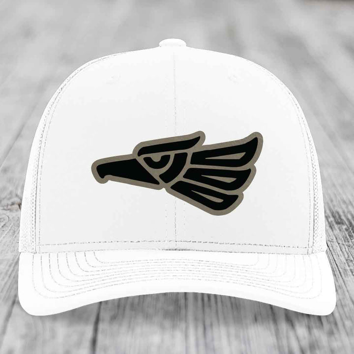 White trucker hat with black and grey Aztec-style eagle patch on front.