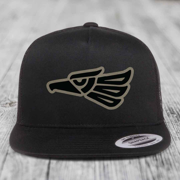Black snapback hat with Aztec-style eagle patch on front, displayed on wooden surface.
