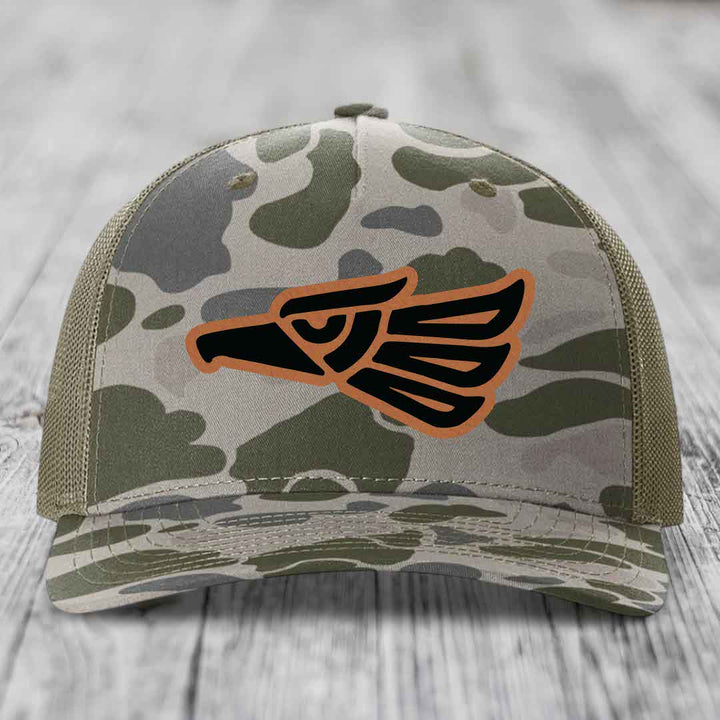 Camouflage trucker hat with black and tan Aztec eagle patch on front.