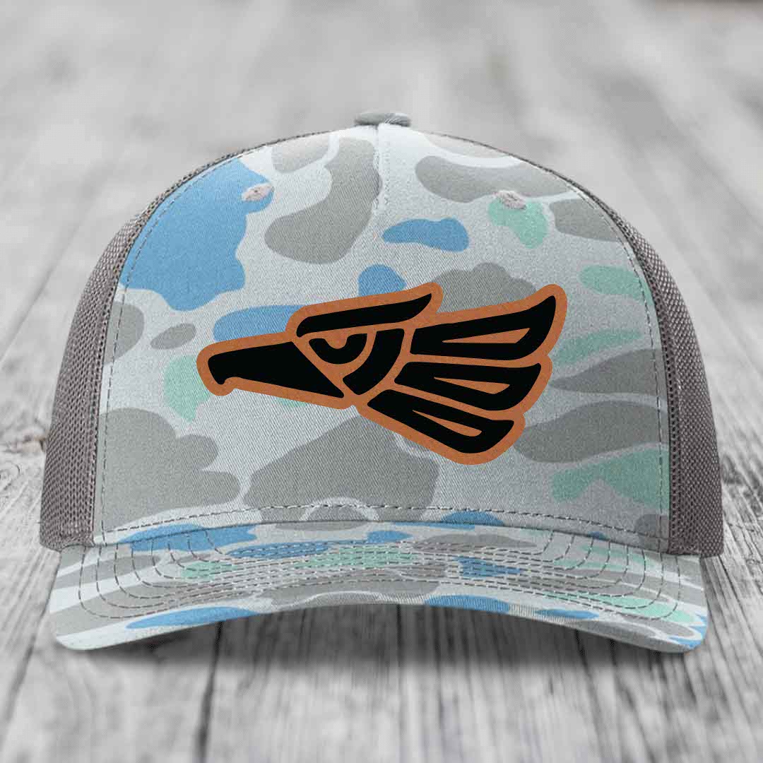 Camouflage trucker hat with Aztec-style eagle patch in black and tan, on wooden surface.