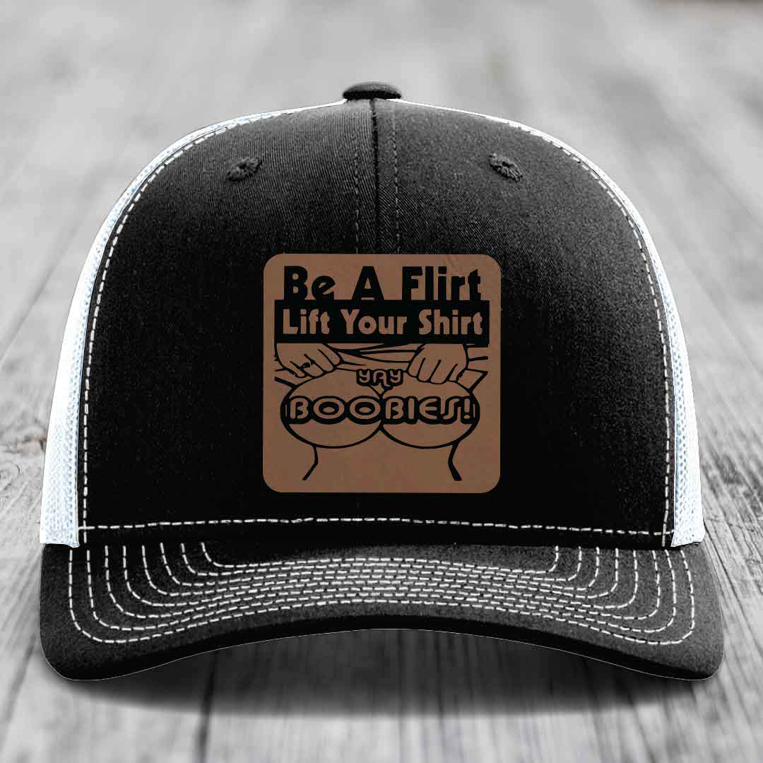 Black trucker hat with white mesh back and stitching, featuring a brown leatherette patch that reads &