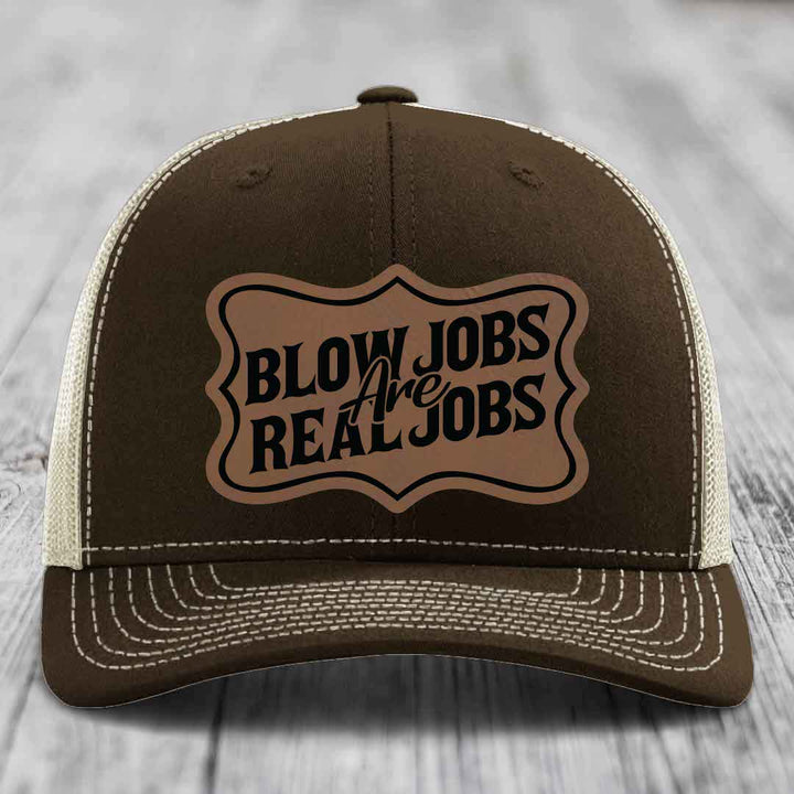 Brown trucker hat with cream mesh back and white stitching, featuring a tan leatherette patch that reads &