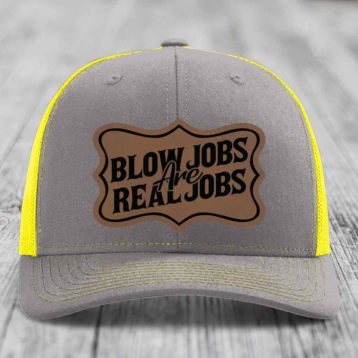 Grey trucker hat with yellow mesh and stitching, featuring a brown leatherette patch that reads &