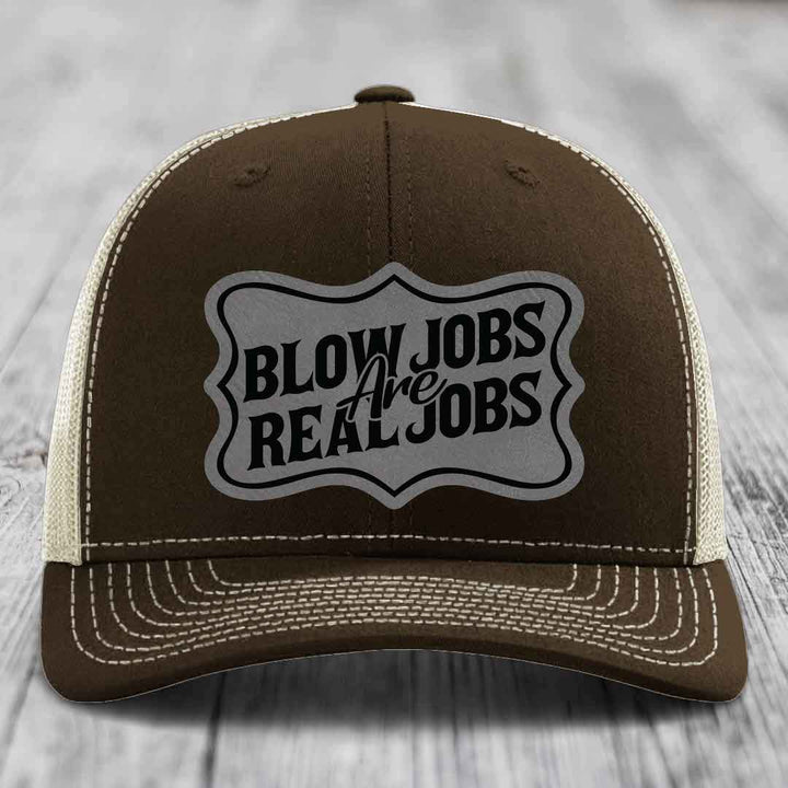 Brown trucker hat with cream mesh back and white stitching, featuring a grey shield patch with black text reading &