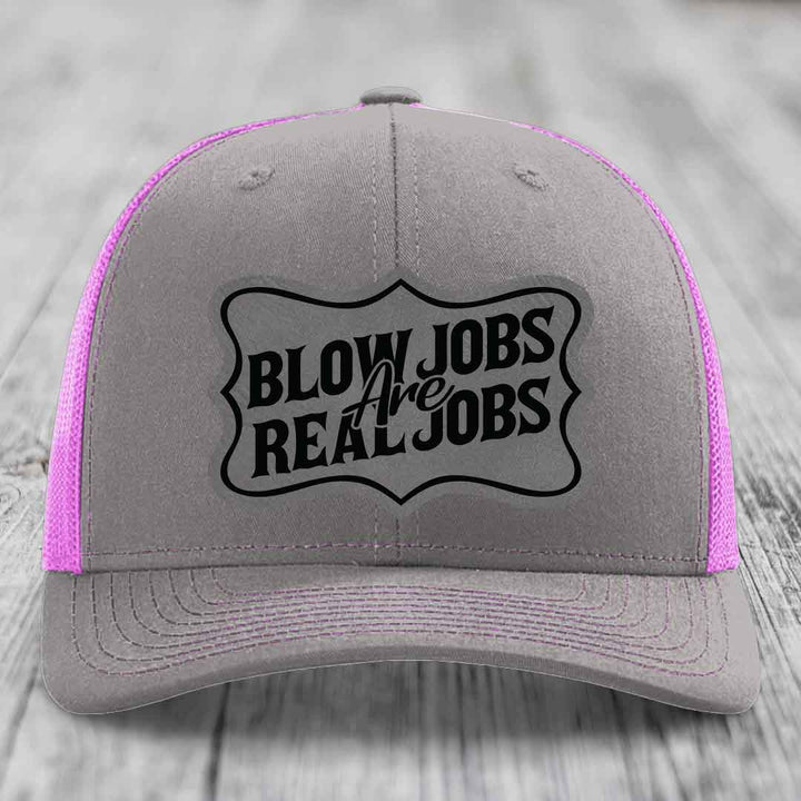 Grey trucker hat with pink mesh sides and black "Blow Jobs Are Real Jobs" patch on front.