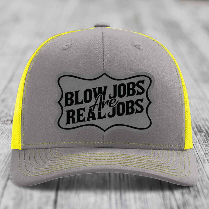 Grey trucker hat with yellow mesh sides and stitching, featuring a black "Blow Jobs Are Real Jobs" patch.