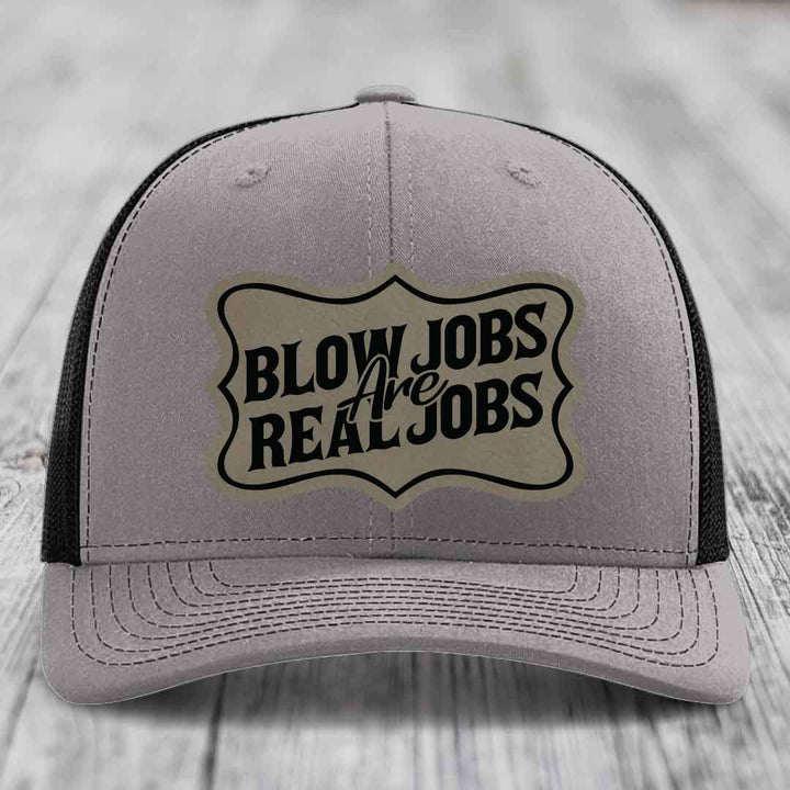 Grey trucker hat with black mesh back and a beige leatherette patch that reads &
