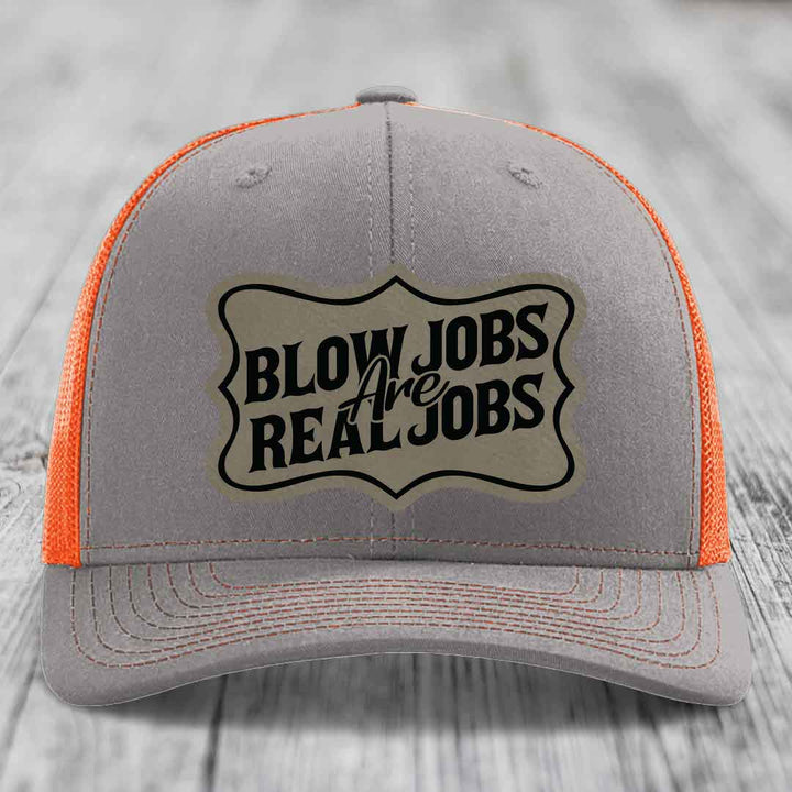 Grey trucker hat with orange mesh back and black embroidered patch reading &