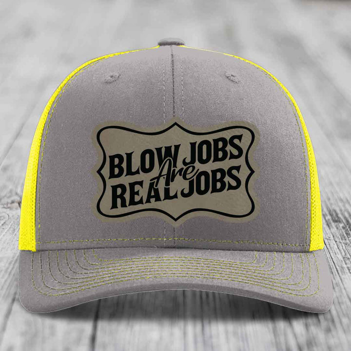 Grey trucker hat with yellow mesh and stitching, featuring a beige leatherette patch that reads &