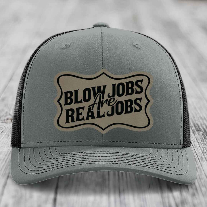 Grey trucker hat with black mesh back and a beige leatherette patch that reads &