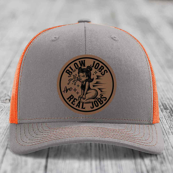Grey trucker hat with orange mesh back and a brown leatherette patch reading &