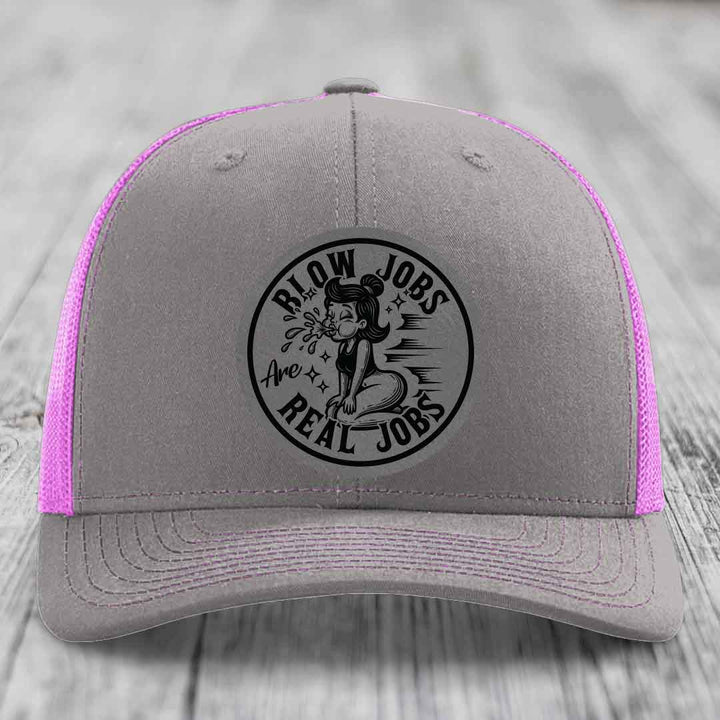 Grey trucker hat with pink mesh back and black circular patch reading &