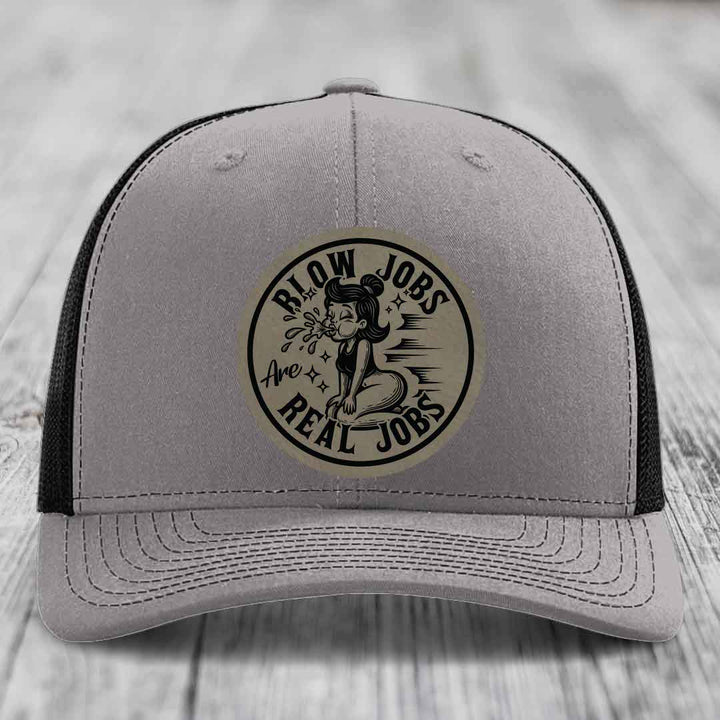 Grey trucker hat with black mesh back and a circular beige leatherette patch reading &