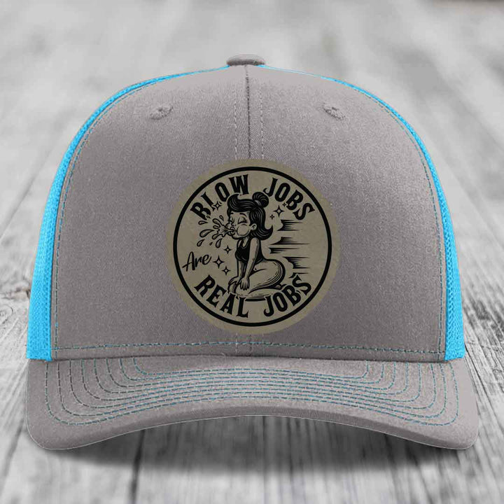Grey trucker hat with blue mesh sides and a circular beige leather patch featuring a cartoon woman and the text &