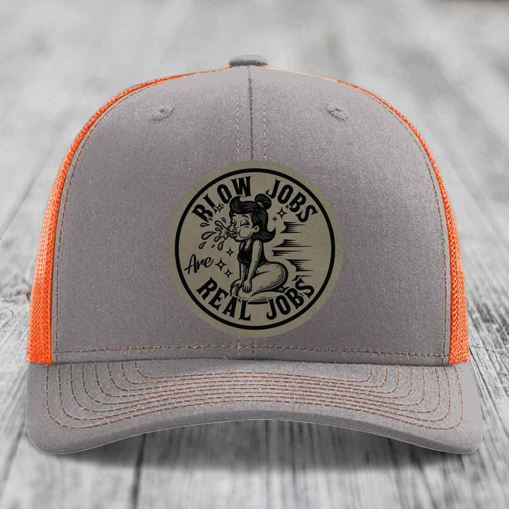 Grey trucker hat with orange mesh back and a circular leatherette patch reading &