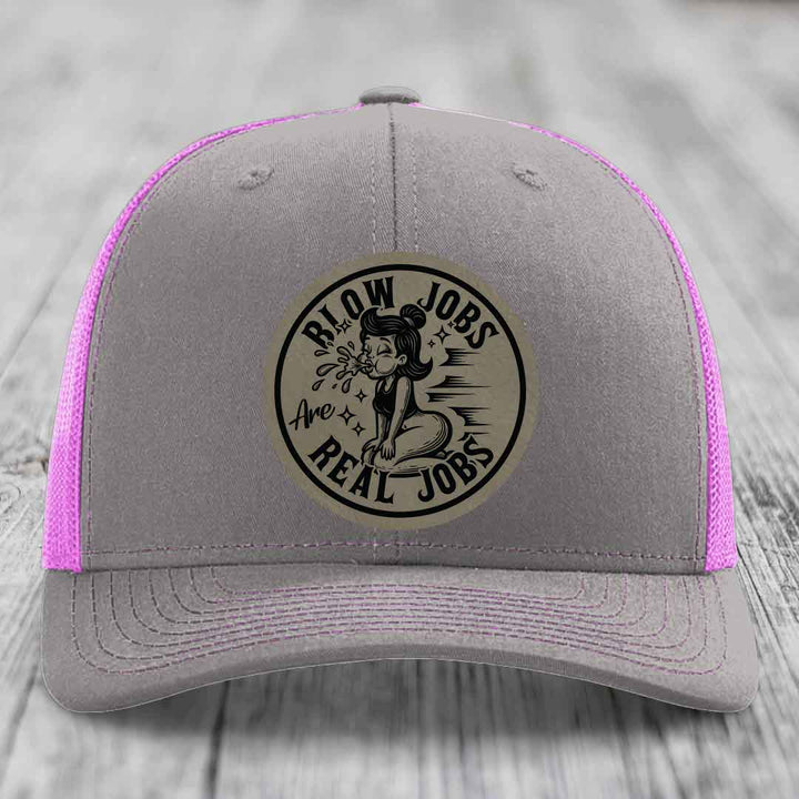 Grey trucker hat with pink mesh sides and a circular leatherette patch reading &
