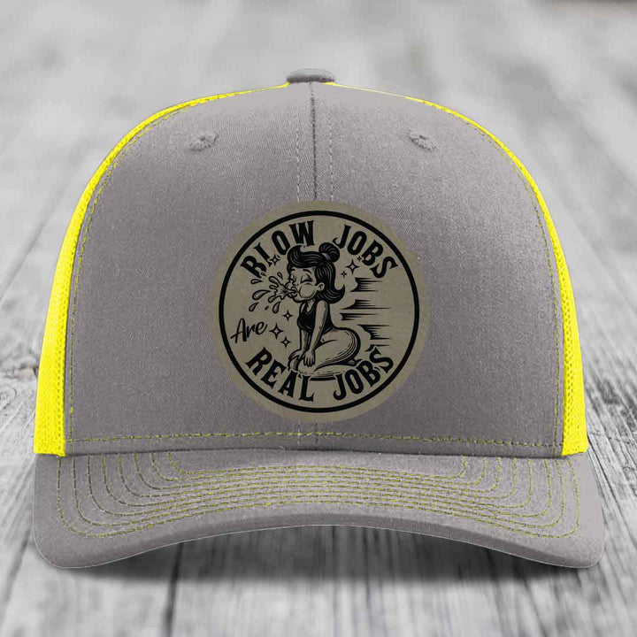 Grey trucker hat with yellow mesh and stitching, featuring a circular patch that reads &