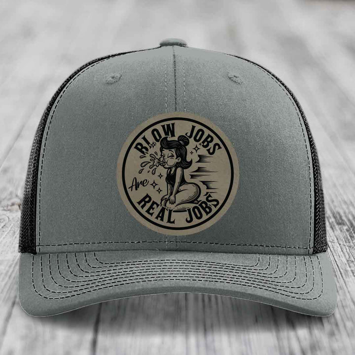 Grey trucker hat with black mesh back and a circular beige leatherette patch featuring a cartoon woman blowing bubbles and the text &