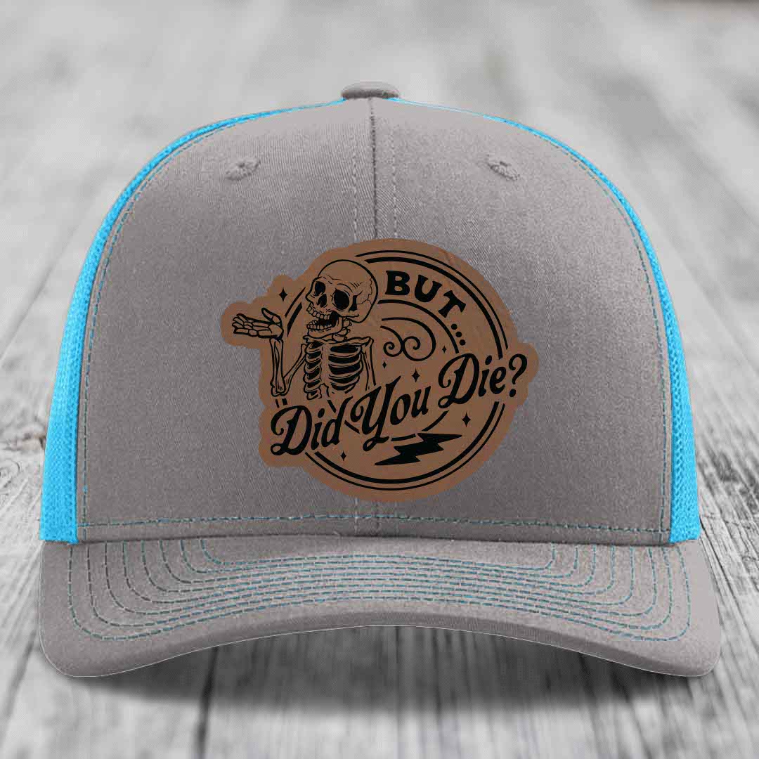 Grey trucker hat with blue mesh sides and a brown leatherette patch featuring a skeleton and text &
