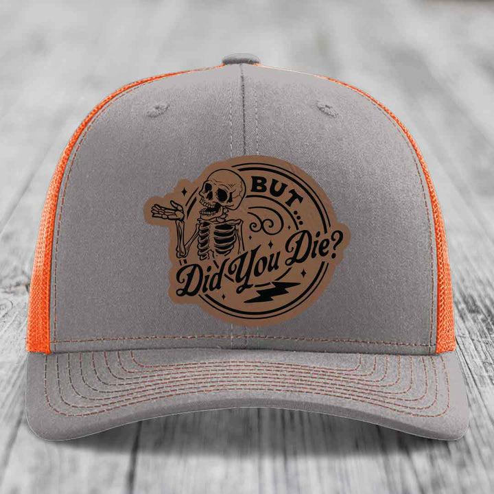 Grey trucker hat with orange mesh back and a brown leather patch featuring a skeleton and text &