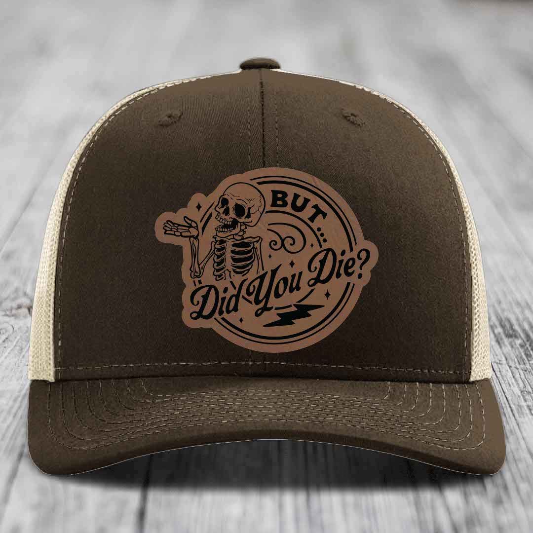 Brown trucker hat with beige mesh back and a circular leather patch featuring a skeleton and text &