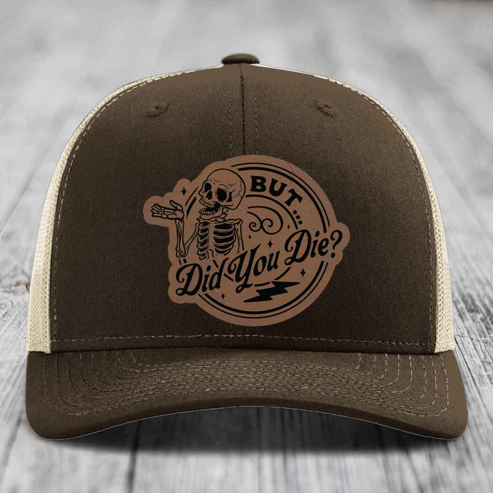 Brown trucker hat with beige mesh back and a circular leather patch featuring a skeleton and text &