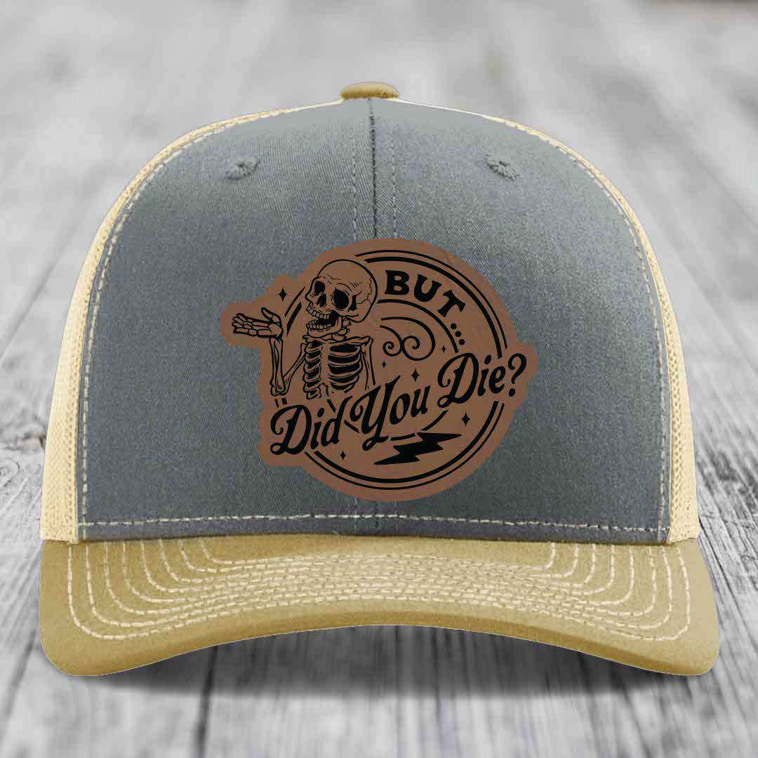 Grey and beige trucker hat with a brown leather patch featuring a skeleton and the text &