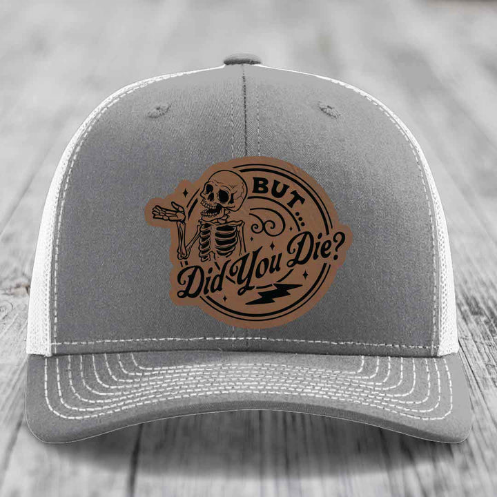 Grey trucker hat with white mesh back and a brown leather patch featuring a skeleton and the text &