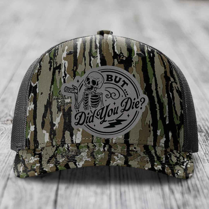 Camouflage trucker hat with a grey circular patch featuring a skeleton and the text &