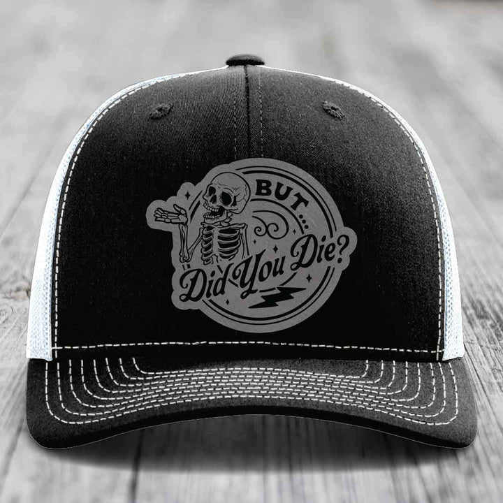 Black trucker hat with white mesh back and stitching, featuring a gray circular patch with a skeleton and text &