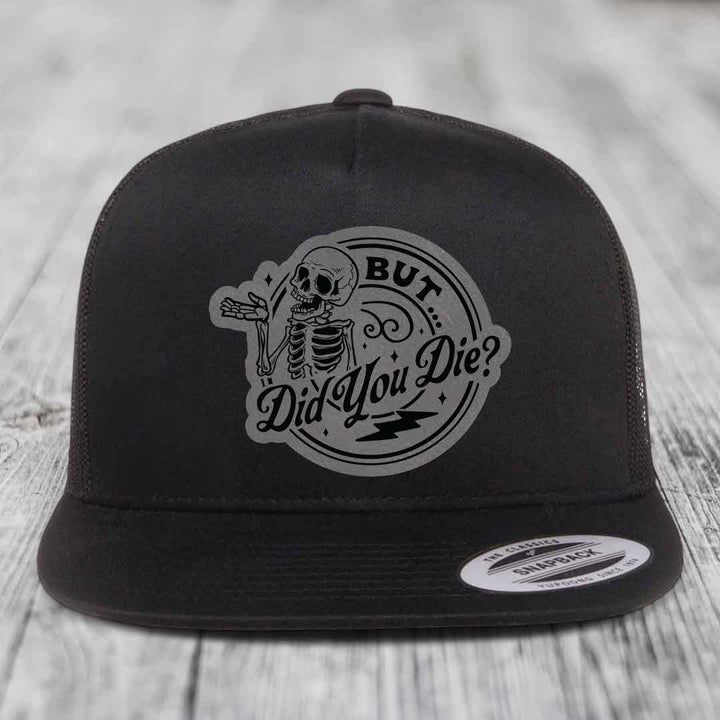 Black snapback hat with a grey circular patch featuring a skeleton and the text &