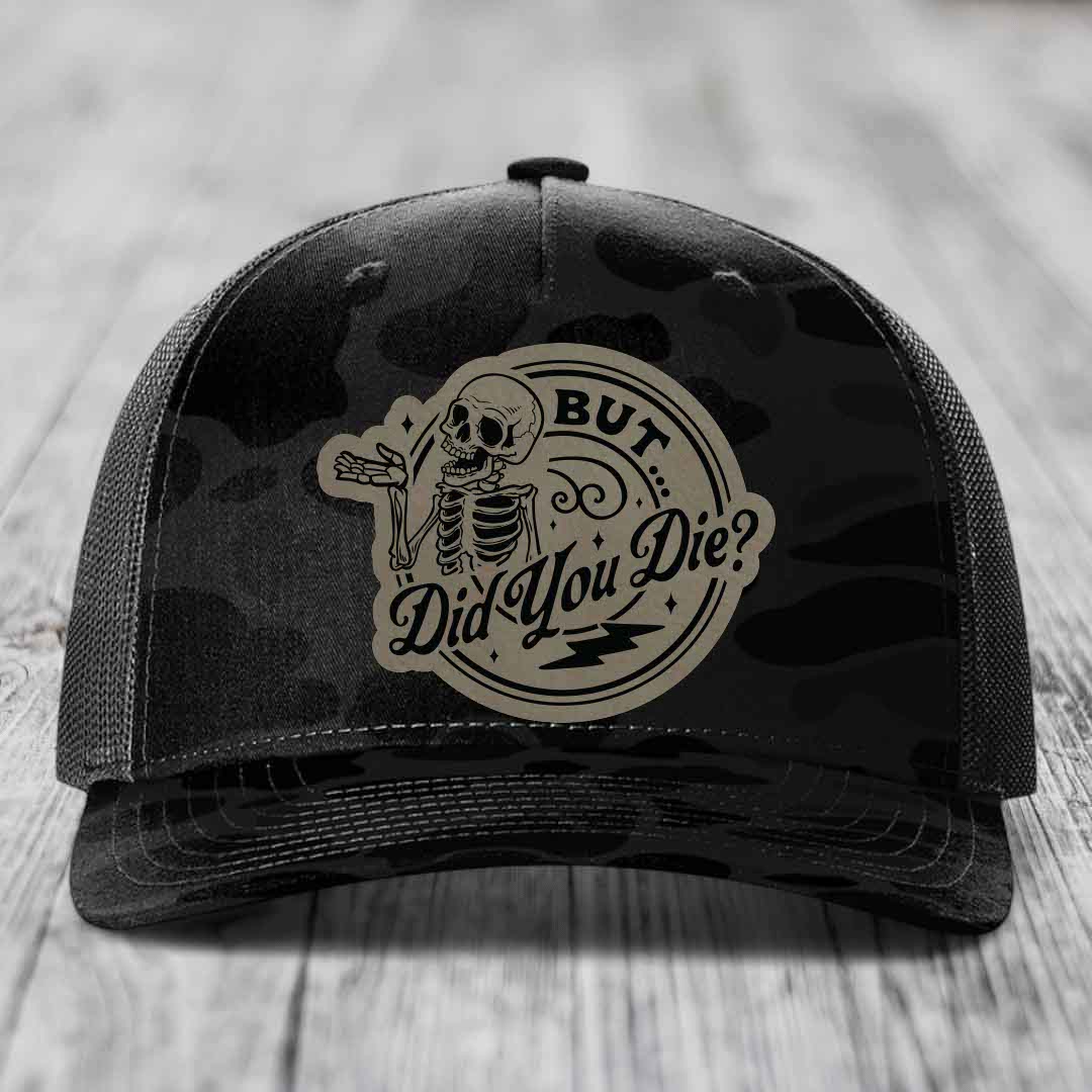 Black camouflage trucker hat with a beige circular patch featuring a skeleton and the text &